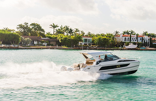 An Aquila 36 power catamaran cruising through clear blue water from left to right with palm trees and houses in the background