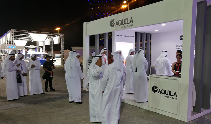 people standing outside of an aquila booth at the abu dhabi international boat show at night